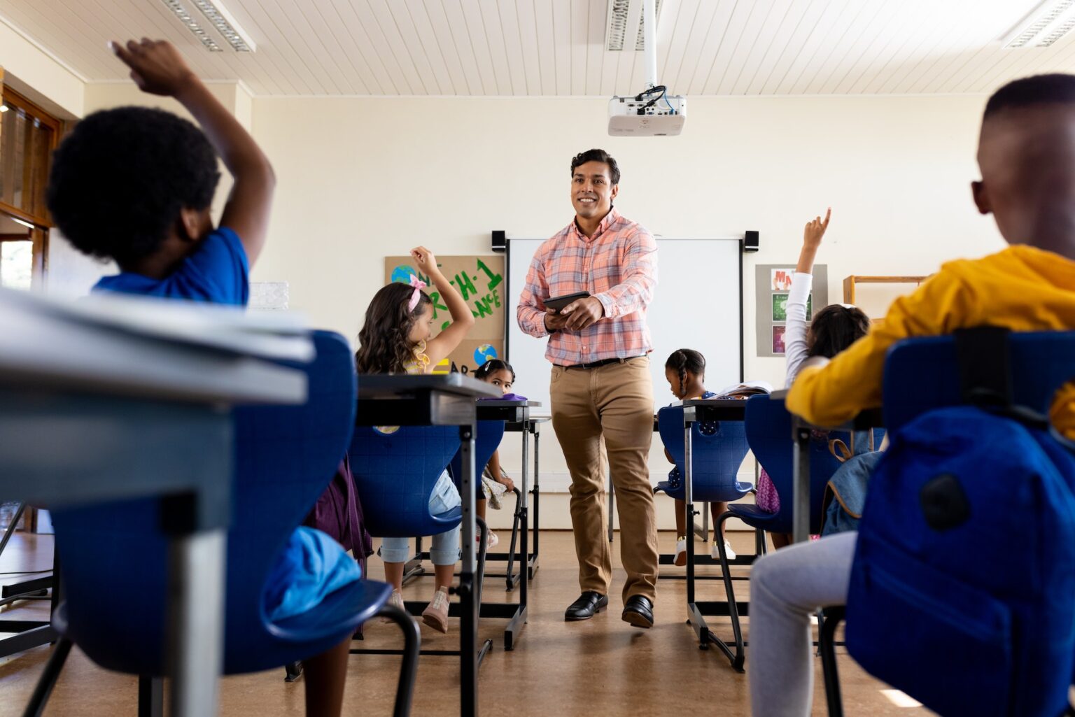 Diverse male teacher with tablet and elementary schoolchildren raising hands in class